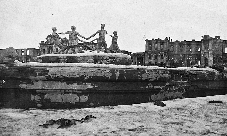 A damaged statue of children holding hands in front of a damaged building